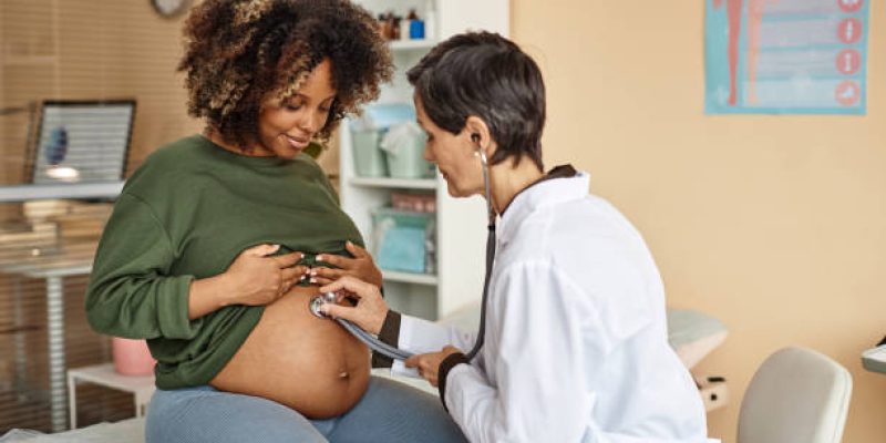 Smiling young African American expectant mother sitting on medical table in clinic while female obstetrician using stethoscope listening baby